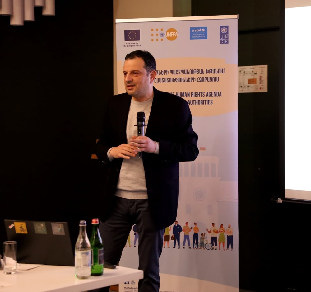 Person presenting at a conference with banners displaying logos of UNICEF, UNDP, EU, and UNFPA in the background.