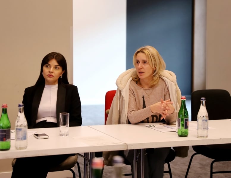 Two women seated at a conference table, listening attentively. One is gesturing while speaking. On the table are water bottles and notepads.