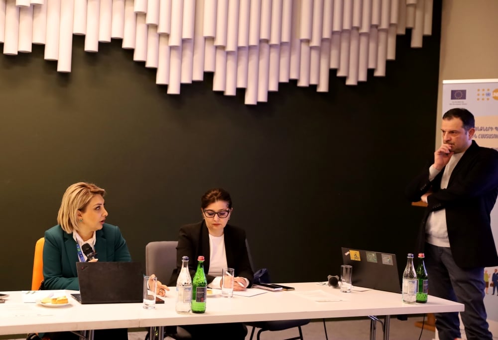 Three participants at a conference table with laptops and water bottles, listening attentively to a speaker in a room with modern lighting fixtures and a banner displaying logos.