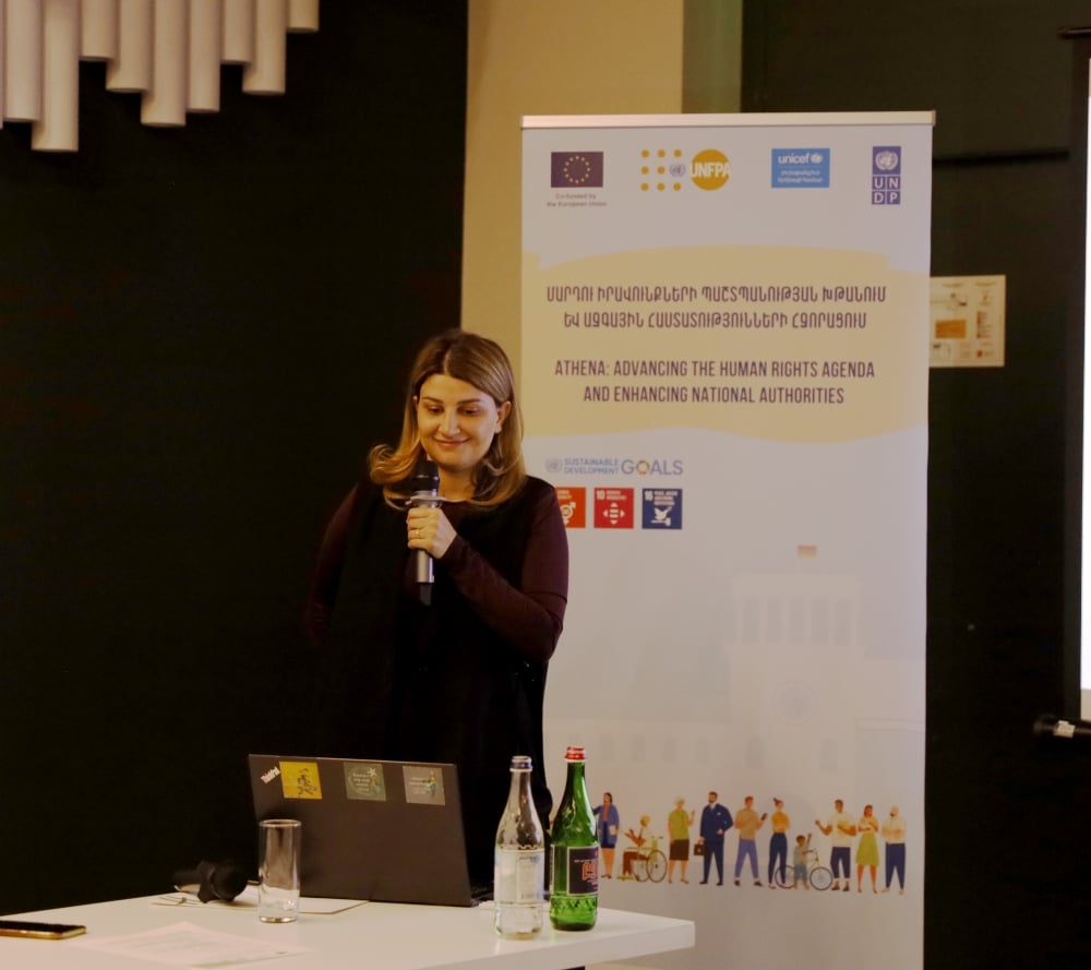 A woman standing at a podium with a laptop, delivering a presentation in a room with banners featuring logos of the European Union, UNICEF, UNFPA and UNDP. 