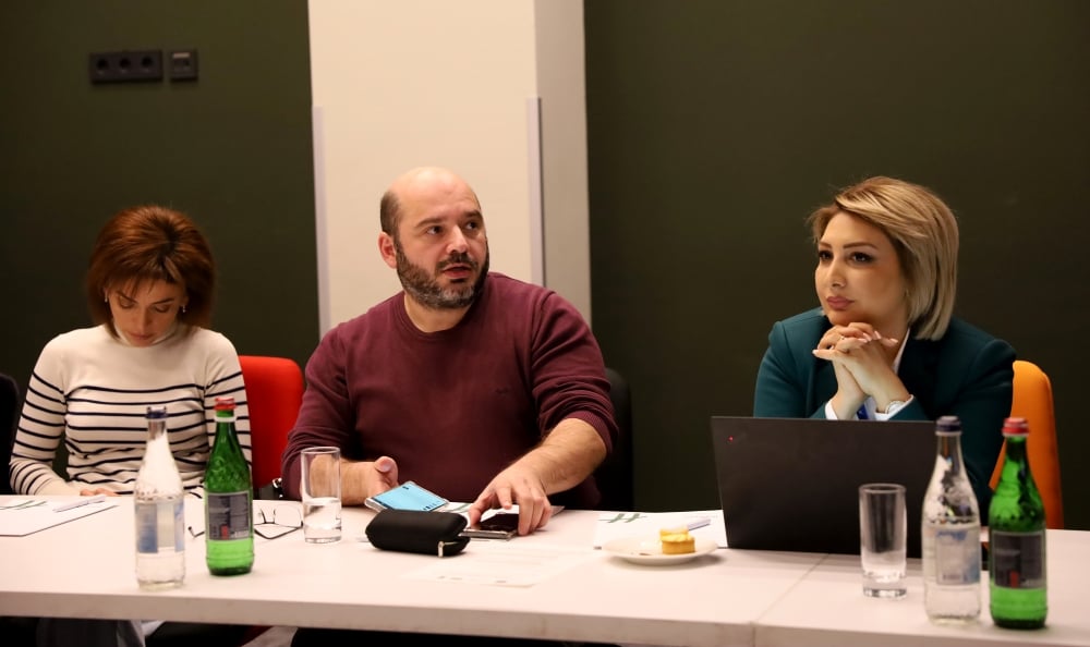 Three professionals are sitting at a table during a meeting with laptops, papers. The man in the center is speaking.