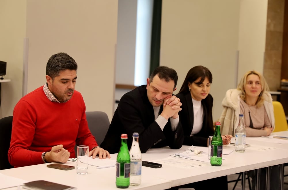 Four professionals seated at a conference table engaged in a discussion. Bottles of water are placed in front of them. All are attentively listening to the individual on the left who is speaking.