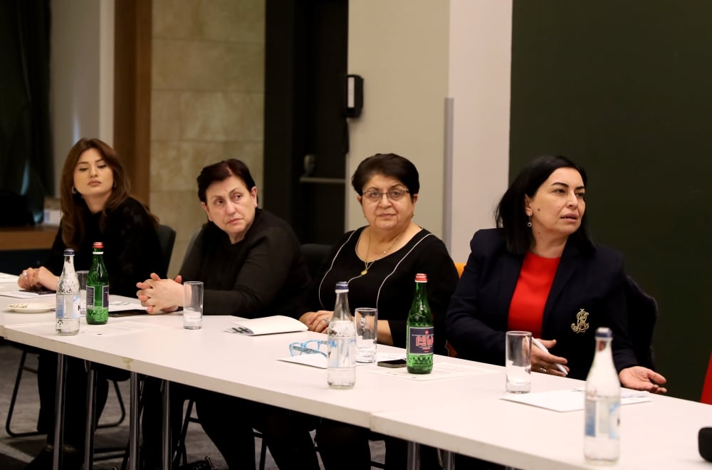 Four women are seated at a conference table with microphones and water bottles,one of them is speaking, the rest are listening. 