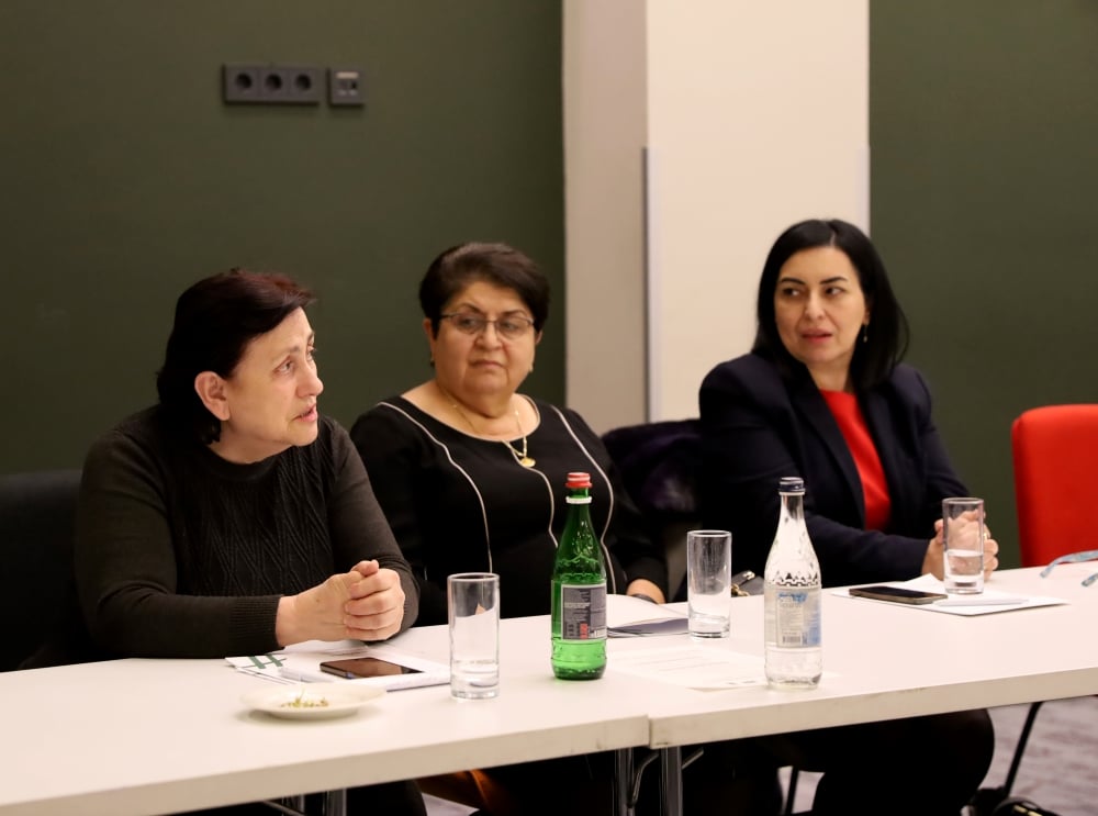 Three women sitting at a conference table during a meeting with water bottles on the table, engaging in a discussion.