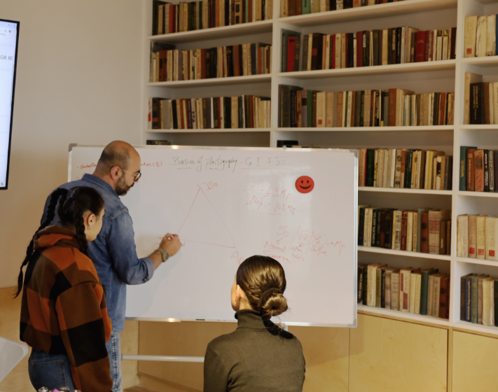 Three people are collaborating around a whiteboard filled with various diagrams and notes, in a room lined with bookshelves.