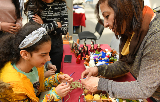 A young girl is in front of a woman teaching them new handicraft skills. She appears to be fully engaged and enthusiastic.