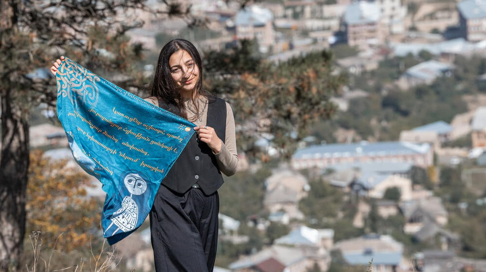 A girl holding a blue scarf, standing outdoors with a scenic background of trees and distant buildings.