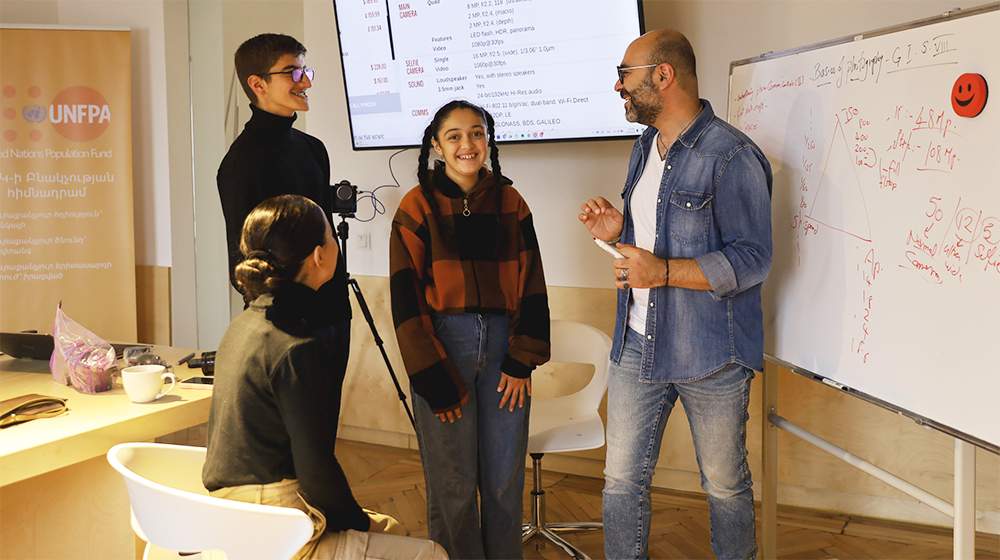 Four people engaging in a lively conversation in a workshop setting with a whiteboard filled with notes and a projected UNFPA presentation in the background.