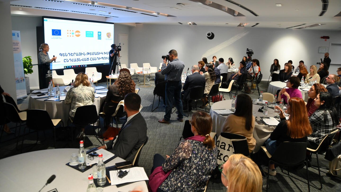 Attendees seated at a conference listening to a speaker at a podium, with cameras filming and event logos of various organizations displayed in the background.