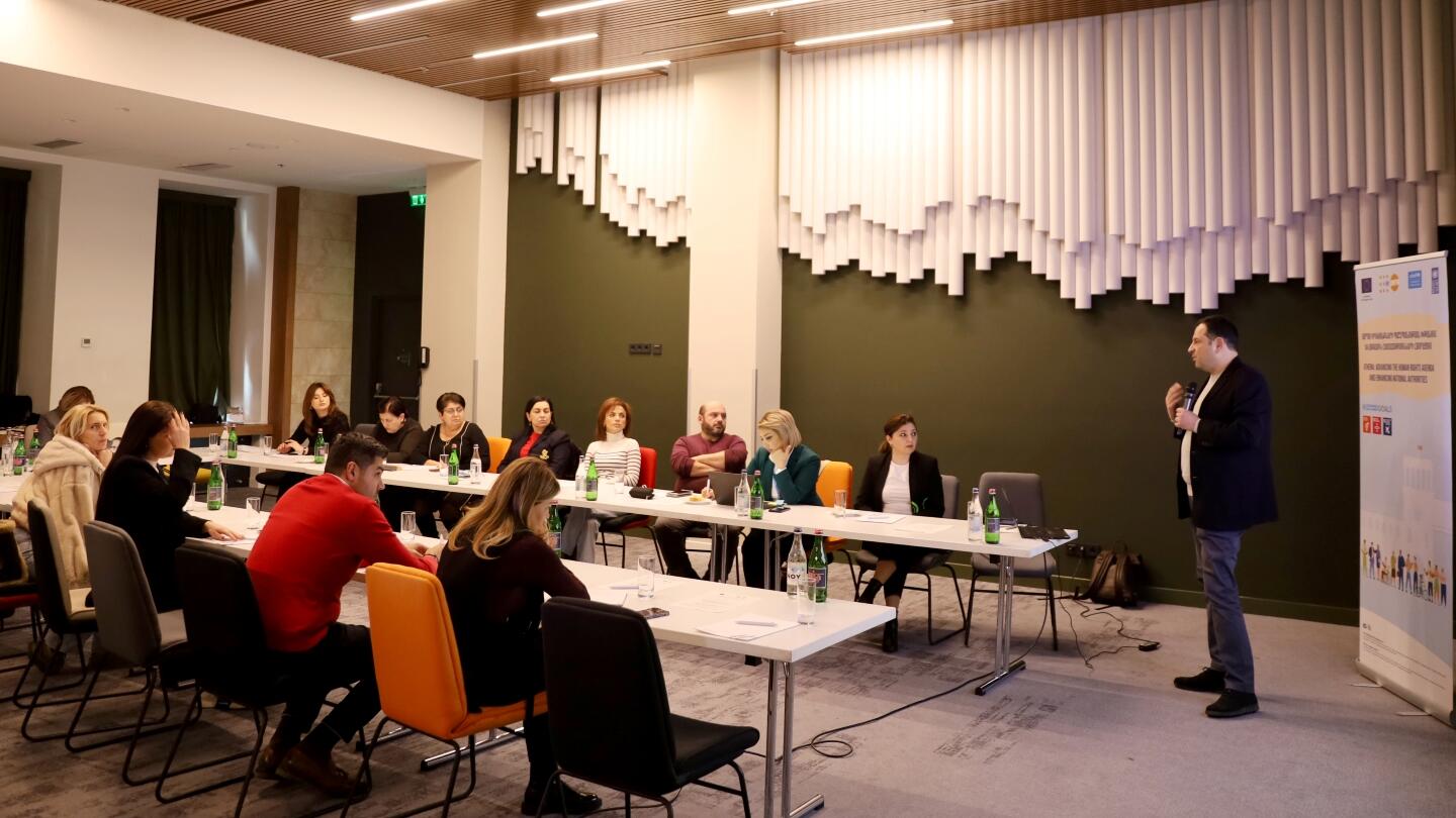 A man presenting at a workshop in a modern conference room with attendees seated around tables listening attentively.