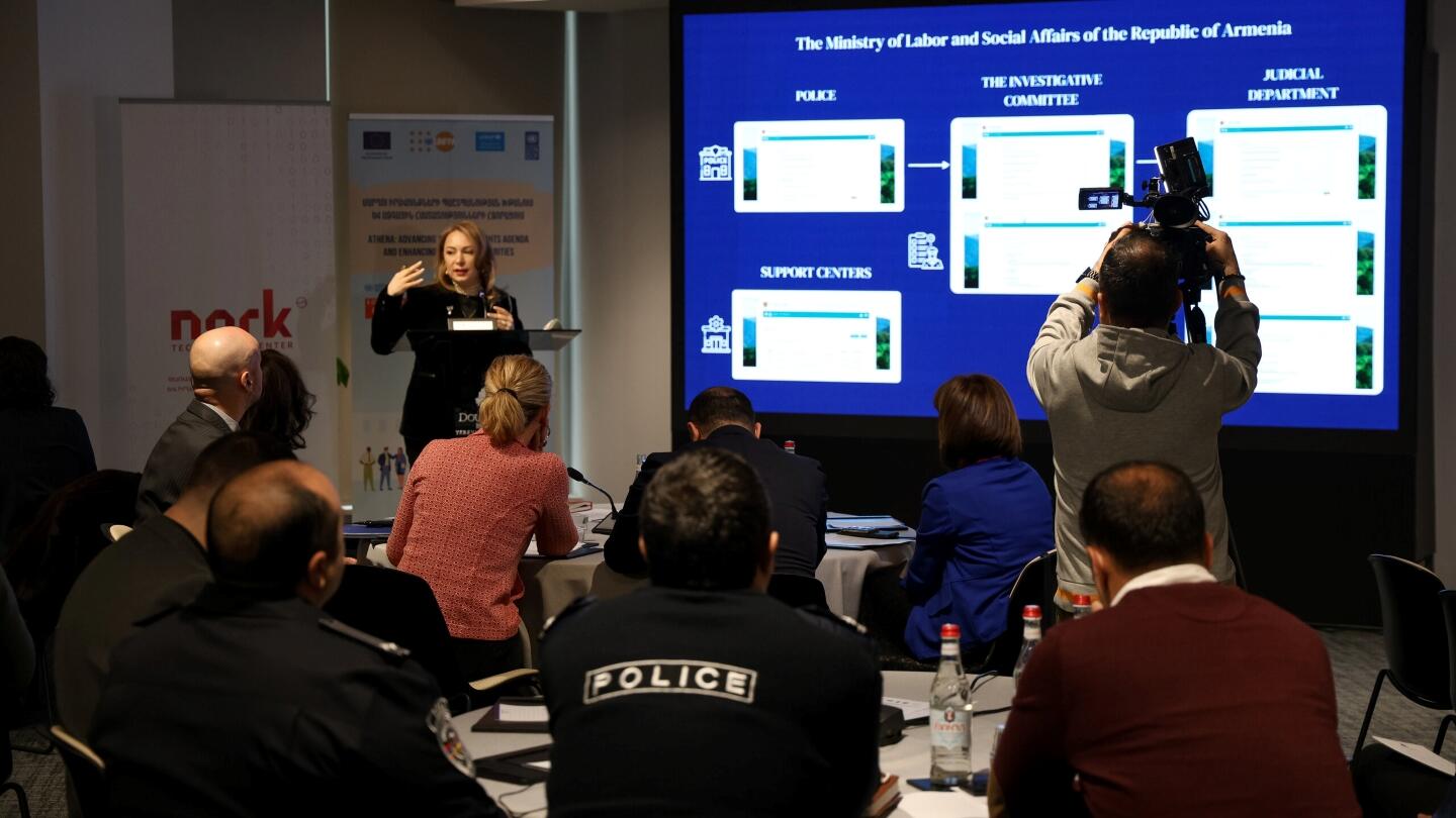 A person presenting at a conference in front of a poster titled "The Ministry of Labor and Social Affairs of the Republic of Armenia" with attendees in uniforms, some visibly labeled "POLICE." One attendee is capturing the event on a video camera.