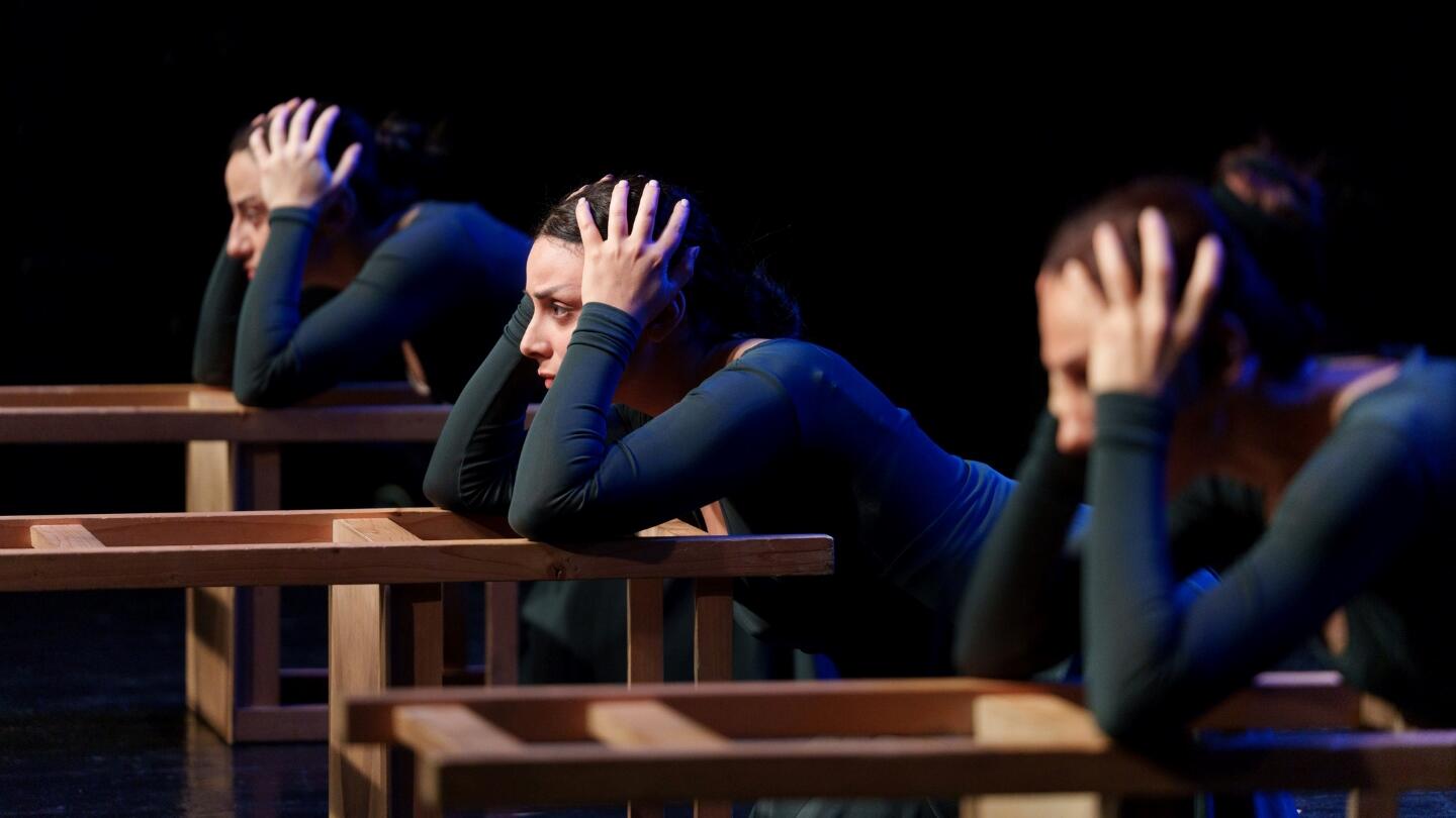 Three performers in blue dresses are seated at wooden benches, each holding their head in a dramatic pose, on a dimly lit stage.