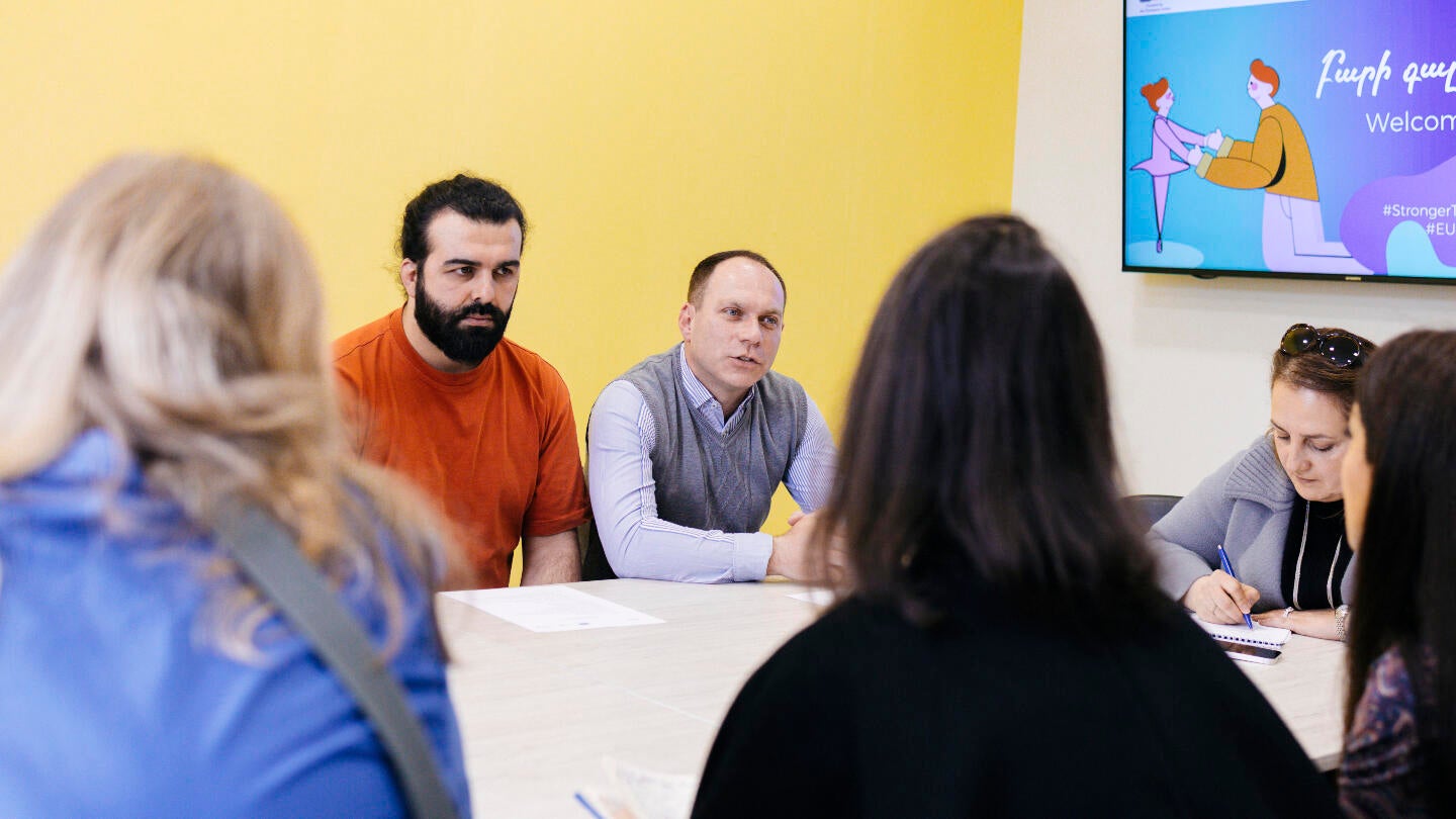Group of people sitting at a table during a meeting with a presentation screen in the background.