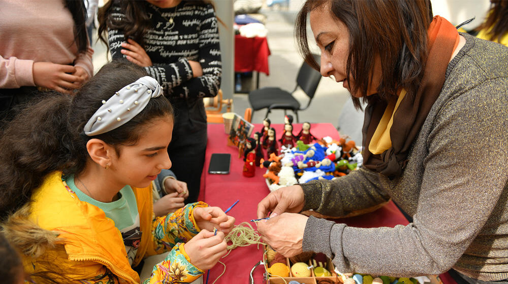 A young girl is in front of a woman teaching them new handicraft skills. She appears to be fully engaged and enthusiastic.