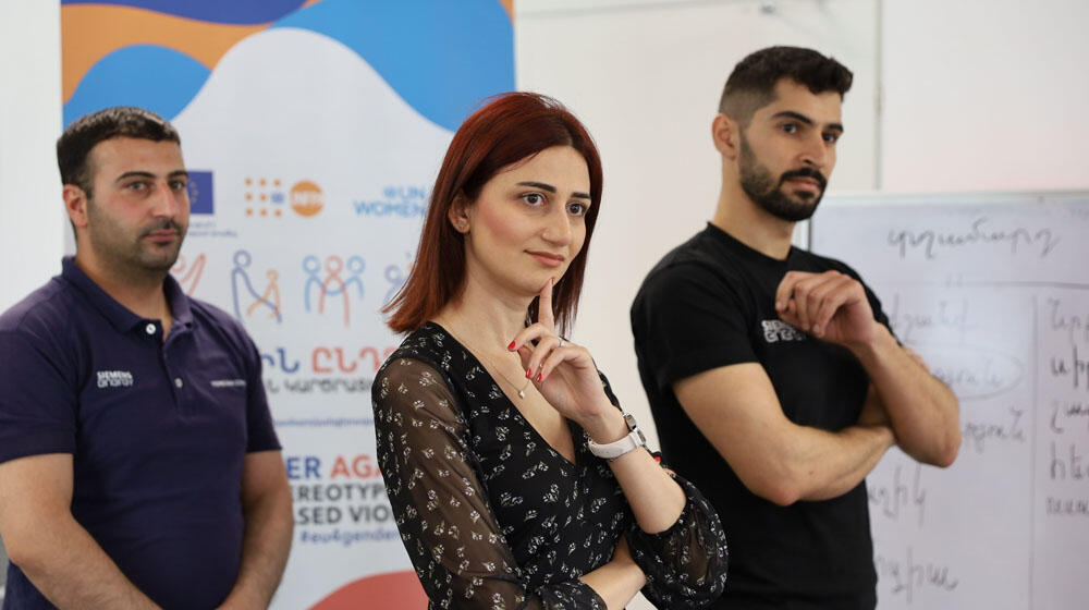 Three young adults, two men and a woman, workshop participants, stand in front of a banner that reads: 'Together against gender 